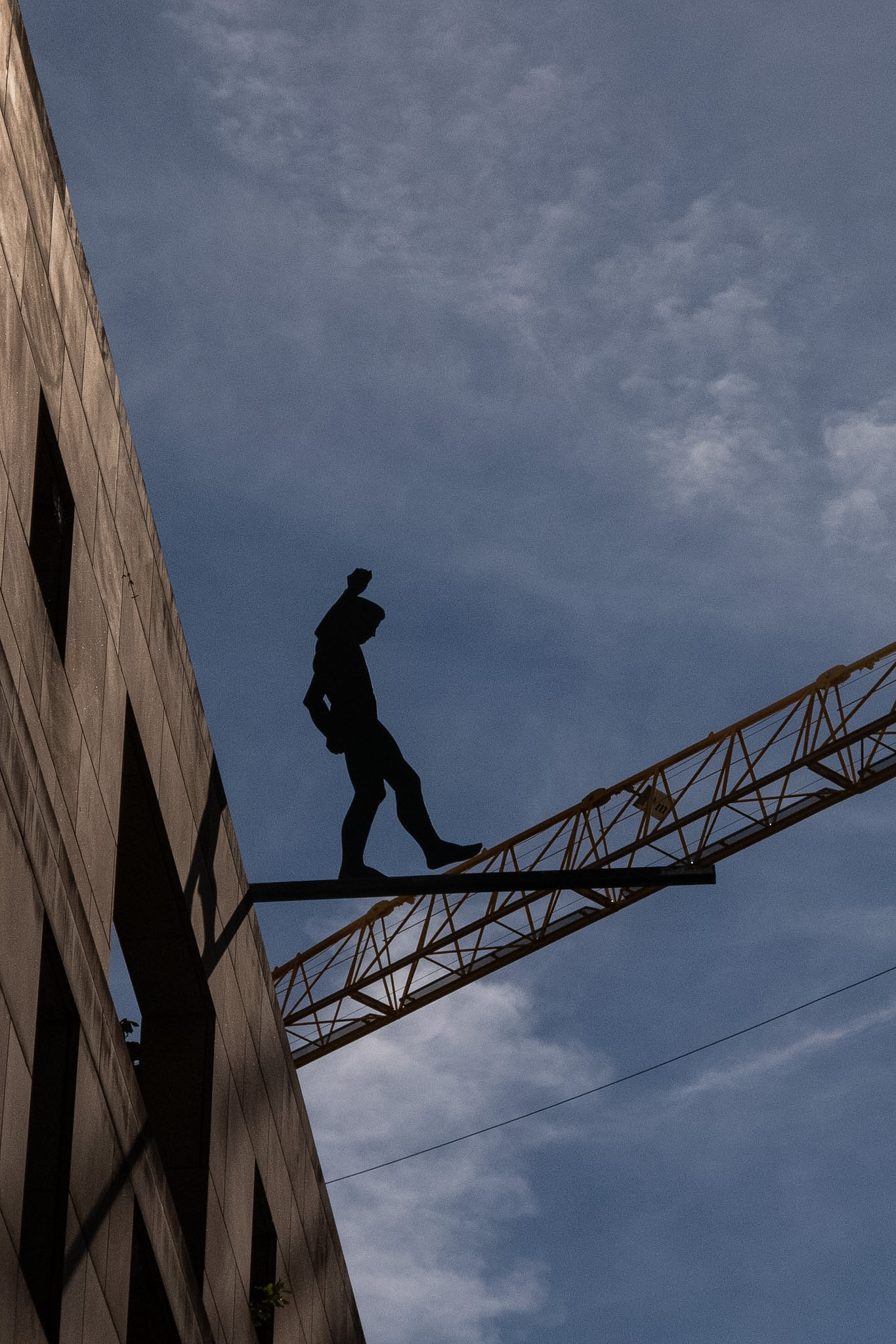 Silhouetted figure on a beam beside a building, with a construction crane against the blue sky